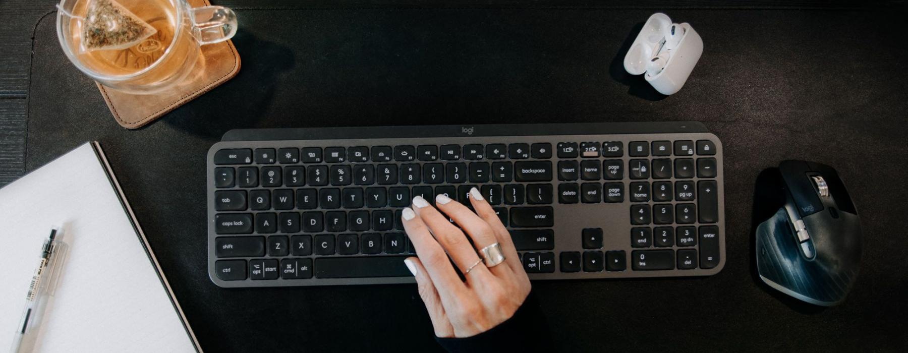 woman's hand on a keyboard surrounded by office items and a cup of tea on a coaster